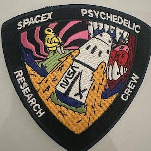 Peut inclure: Un écusson triangulaire noir avec les mots "SpaceX" et "Psychedelic Crew" en haut et en bas, respectivement. L'écusson présente un design psychédélique coloré avec une fusée, une créature aux dents et une figure jaune.