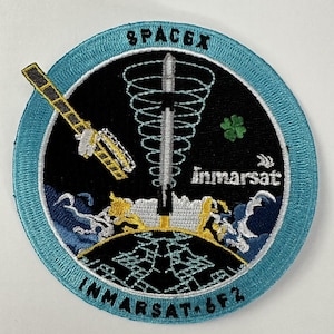 May include: A round, blue and black embroidered patch with the text "SPACEX" and "INMARSAT, 6F2" around the edge. The centre of the patch features a white rocket with a green shamrock, a satellite, and a blue spiral.