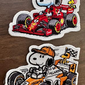 Op de afbeelding: Twee geborduurde patches met Snoopy die in raceauto's rijdt. Eén patch toont Snoopy in een rode Ferrari raceauto, en de andere toont Snoopy in een oranje McLaren raceauto. Beide patches hebben een witte rand.