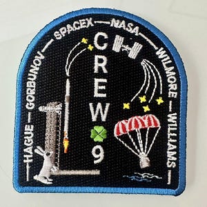 May include: A black and white embroidered patch with a blue border. The patch features the text "CREW 9" and the names "HAGUE - GORBUNOV - SPACEX - NASA - WILMORE - WILLIAMS". The patch also includes a rocket, a rabbit, a four-leaf clover, and a parachute.