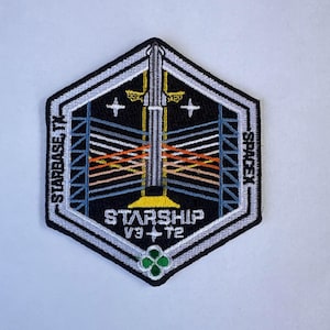 Peut inclure: Un écusson brodé hexagonal présentant une fusée argentée centrale sur fond noir. Le motif inclut des structures de tour de lancement bleues, des lignes horizontales orange et blanches, et le texte STARBASE, TX, SPACEX et STARSHIP V3+T2.