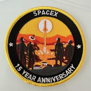May include: A round, yellow-bordered patch with a black background. The patch features a white rocket launching on a red and orange landscape with a white sun. The text "SPACEX" is at the top of the patch, and "15 YEAR ANNIVERSARY" is at the bottom.