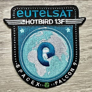 Könnte beinhalten: Ein blauer und weißer gestickter Patch mit dem Text "Eutelsat Hotbird 13F" und einem Globus mit einem blauen "E" in der Mitte. Der Text "SpaceX Falcon 9" ist um den Globus herum geschrieben.