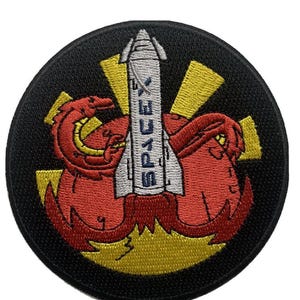 Puede incluir: Un parche bordado negro y amarillo con un dragón rojo envuelto alrededor de un cohete blanco con la palabra "SPACEY" escrita en el lateral. El cohete se lanza desde un planeta rojo con llamas.