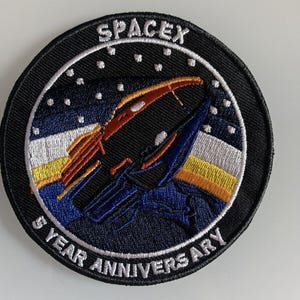Puede incluir: Un parche negro circular con el texto "SPACEX" en la parte superior y "5 YEAR ANNIVERSARY" en la parte inferior. El parche presenta un diseño de cohete en azul, naranja y negro sobre un fondo azul estrellado.