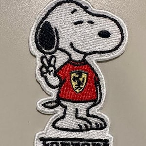 Puede incluir: Parche bordado con Snoopy vistiendo una camiseta roja de Ferrari, haciendo el signo de la paz. El parche es blanco con detalles en negro y la palabra "ferrari" en la parte inferior. Un artículo decorativo para ropa o accesorios.