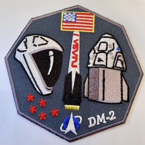 以下が含まれることがあります： NASAのロケット、宇宙飛行士のヘルメット、宇宙船が刺繍されたパッチ。パッチには、アメリカ国旗、赤い星、テキスト「NASA」と「DM-2」が、濃い青色の八角形の背景に描かれています。