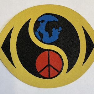 Op de afbeelding: Een gele patch met een zwart yin-yang ontwerp. De bovenste helft heeft een blauwe wereldbol en de onderste helft een rood vredesteken. De patch is geel omlijnd.
