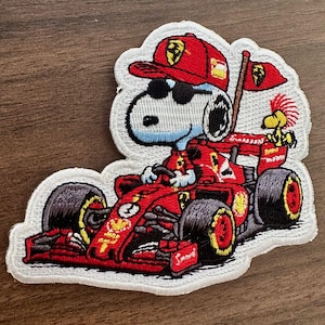 May include: Embroidered patch featuring Snoopy in a red Ferrari race car, wearing a red cap and sunglasses. The patch includes a flag and Woodstock. The patch has a white border and is on a wooden surface.