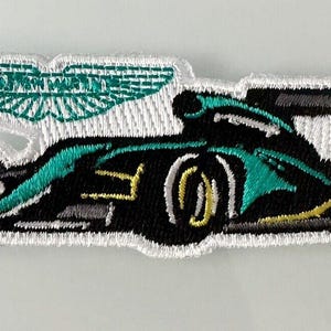 May include: Embroidered patch of a Formula 1 race car in black, teal, and yellow. The Aston Martin logo is above the car. The patch has a white border.