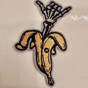 May include: Embroidered patch featuring a yellow banana with a black outline, peeled open to reveal a skeleton hand in a shaka gesture. The patch has a gray border and is likely for clothing or accessories.