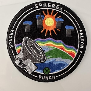 May include: A black and white embroidered patch with the text "SPHEREX - SPACEX - FALCON 9 - PUNCH" around the edge. The center of the patch features a stylized image of a satellite, a rainbow, a sun, and a planet.