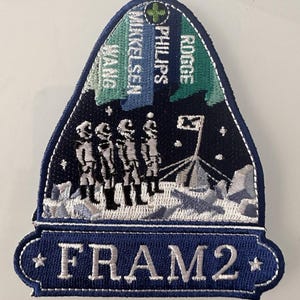 May include: A blue and white embroidered patch with a moon landing scene and the text "FRAM 2". The patch features three astronauts standing on the moon with a flag planted in the ground. The names "Rodger", "Phillips", "Winkelseen", and "Wang" are embroidered on the top of the patch.