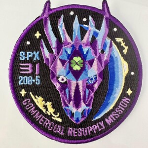Peut inclure: Un écusson brodé violet, bleu et jaune avec un motif de tête de dragon. L'écusson porte le texte "SPX-31 208-5 COMMERCIAL RESUPPLY MISSION" sur le bord.