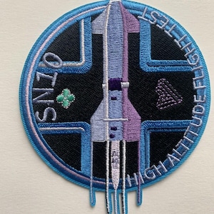 Puede incluir: Un parche bordado azul y blanco con un diseño de cohete. El texto "HIGH ALTITUDE" está escrito alrededor del borde del parche.