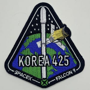 以下が含まれることがあります： 黒色の三角形のワッペンで、白いロケットが青と緑の地球から打ち上げられている。地球には「KOREA 425」という文字が白で刺繍されている。ワッペンの下部には「SPACEX」と「FALCON 9」という文字が白で刺繍されている。