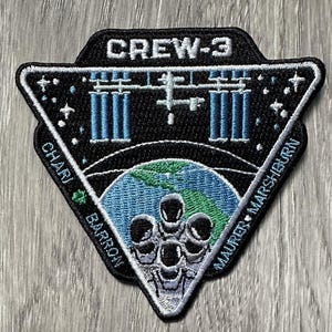 Peut inclure: Un écusson brodé noir et blanc de forme triangulaire. L'écusson présente le texte "CREW-3" en haut, une représentation de la Station spatiale internationale au centre et les noms "CHARI", "BARRON" et "MAURER, MARSHBURN" sur les côtés. L'écusson comprend également un globe terrestre bleu et vert avec un groupe d'astronautes en blanc.