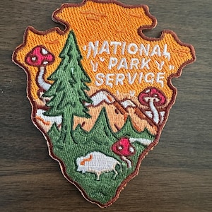 May include: Embroidered patch in the shape of an arrowhead, featuring the text "NATIONAL PARK SERVICE". The patch has a vibrant orange border and background, with a green tree, mountains, mushrooms, and a bison. A colorful outdoor-themed patch.
