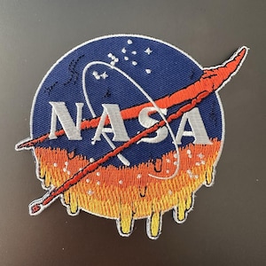 Può includere: Toppa ricamata con il logo NASA e un design a fusione. La toppa ha uno sfondo blu con testo bianco e uno swoosh rosso. La parte inferiore della toppa ha un effetto di fusione con colori arancioni e gialli.