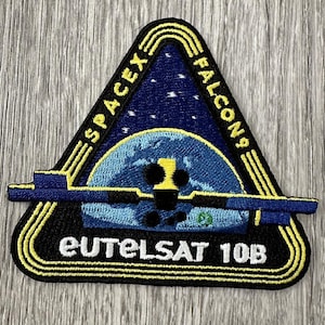 May include: A SpaceX Falcons Eutelsat 10B patch. The patch is triangular with a black border and yellow accents. The design features a blue and white Earth, a rocket, and the text "SPACEX FALCON 9" and "EUTELSAT 10B".
