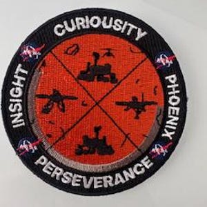 Könnte beinhalten: Ein runder, schwarz-orangener gestickter Aufnäher mit den Worten "Curiosity", "Insight", "Perseverance" und "Phoenix" am Rand. Die Mitte des Aufnähers zeigt ein stilisiertes Bild der Mars-Rover Curiosity, Perseverance und Insight sowie der Phoenix-Landesonde.