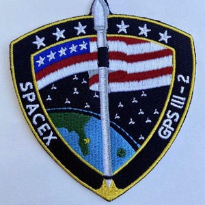 May include: A black and yellow embroidered patch with a white rocket, a US flag, and the text "SPACEX GPS III-2".