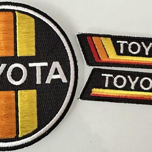 Puede incluir: Tres parches bordados con la palabra "TOYOTA" en letras blancas sobre fondo negro. Los parches tienen rayas rojas, naranjas y amarillas.
