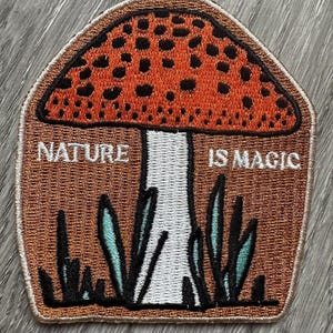 Könnte beinhalten: Ein gestickter Aufnäher mit einem roten und weißen Pilz mit schwarzen Punkten auf dem Hut. Der Pilz ist von grünem Gras umgeben und der Text "NATURE IS MAGIC" steht unter dem Pilz.