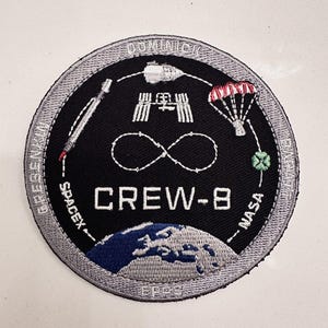 Peut inclure: Écusson brodé noir circulaire avec le texte "CREW-8" et les logos SpaceX et NASA. L'écusson présente des images d'une fusée, d'une station spatiale, d'un parachute et de la Terre. La bordure est grise.