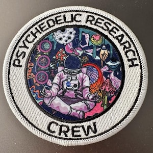 May include: Round embroidered patch with the words "PSYCHEDELIC RESEARCH CREW" in white on a gray border. The patch features an astronaut in a meditative pose surrounded by colorful, surreal imagery.