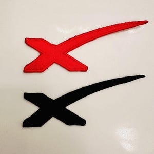 Può includere: Due toppe ricamate a forma di "X". Una toppa è rossa, l'altra nera. Le toppe sono su uno sfondo bianco. La "X" rossa ha una linea più lunga e curva che si estende dal lato destro.