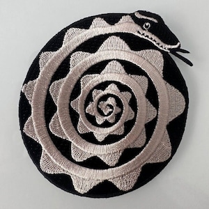 Op de afbeelding: Een geborduurde patch in zwart en wit van een slang die in een spiraalvorm is opgerold.