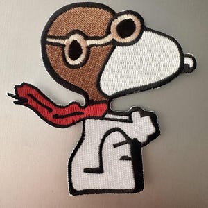 May include: Embroidered patch featuring Snoopy as a World War I flying ace. The patch shows Snoopy in a brown aviator helmet and goggles, a white shirt, and a red scarf. The patch is outlined in black.