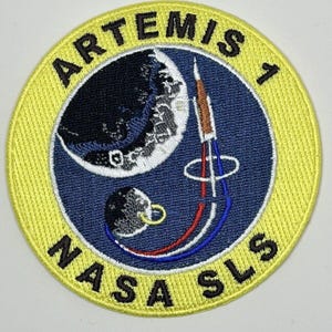 May include: A yellow circular patch with the text "ARTEMIS 1" and "NASA SLS" around a blue background with a white crescent moon, a smaller black sphere, and a red, white, and blue rocket.