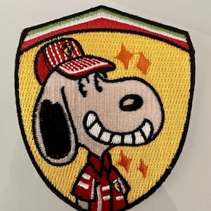 Peut inclure: Écusson brodé représentant Snoopy portant une casquette et une veste rouges et blanches, sur fond jaune et de forme bouclier. L'écusson a une bordure verte, blanche et rouge, et des étoiles orange.