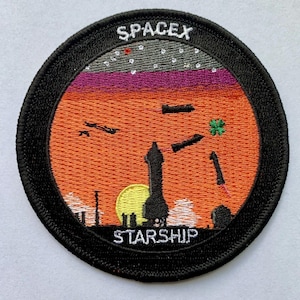 Puede incluir: Un parche bordado en blanco y negro con las palabras "SpaceX" y "Starship" en blanco. El parche representa un cohete que se lanza hacia un cielo de puesta de sol con un sol amarillo y un trébol verde.