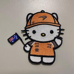 May include: Embroidered patch featuring a cartoon cat character wearing an orange uniform and hard hat. The character is holding a small flag with a blue background and white stars. The patch has black and white details.