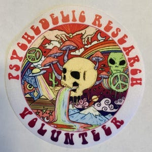 May include: A round patch with the words "PSYCHEDELIC RESEARCH VOLUNTEER" in red. The patch features a colorful illustration of a skull, mushrooms, a rainbow, an alien, and a peace sign.