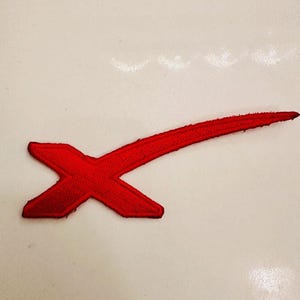 Può includere: Toppa ricamata rossa con una forma a X stilizzata e una linea curva che si estende dall'angolo in alto a destra.