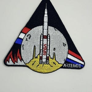 May include: A triangular patch with a white moon and a rocket launching from the surface. The rocket is labeled "NASA" and the moon has a red, white, and blue stripe. The text "ARTEMIS" is on the bottom right corner of the patch.