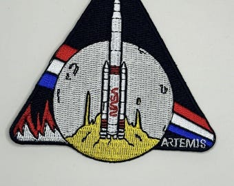 May include: A triangular patch with a white moon and a rocket launching from the surface. The rocket is labeled "NASA" and the moon has a red, white, and blue stripe. The text "ARTEMIS" is on the bottom right corner of the patch.