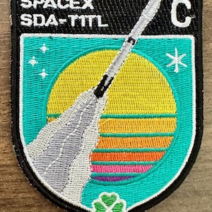 Può includere: Toppa ricamata con un razzo che decolla da un tramonto giallo e arancione, con la scritta "SPACEX SDA-TITL". La toppa ha uno sfondo turchese, un bordo nero e un quadrifoglio in basso.