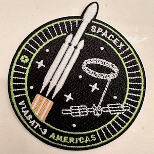 May include: Embroidered patch with a rocket, satellite, and text "SPACEX" and "VIASAT-3 AMERICAS". The design is on a black circular background with a green border. The patch has white and orange accents. A space-themed item.