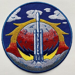 May include: Circular embroidered patch depicting a rocket launch. The design includes a blue sky with white clouds, a yellow and red sun, and a grey and blue foreground. The rocket is white and blue, set against a blue background.