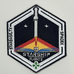 Puede incluir: Parche bordado hexagonal con fondo negro, que presenta un diseño de cohete en amarillo, rojo y azul. El parche incluye el texto "STARBASE TX" y "SPACEX", con "STARSHIP FLIGHT 2" en la parte inferior. También hay un trébol verde.