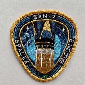 Può includere: Toppa ricamata con il razzo SpaceX Falcon 9. La toppa ha una combinazione di colori blu e oro con il testo "SXM-7" e "SPACEX FALCON 9". Il design include un razzo e delle stelle.