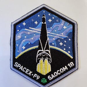 Puede incluir: Parche bordado hexagonal que representa un cohete lanzándose al cielo estrellado. El parche incluye el texto "SPACEX F9" y "SAOCOM 1B". El diseño presenta una combinación de colores azul, negro, amarillo y blanco.
