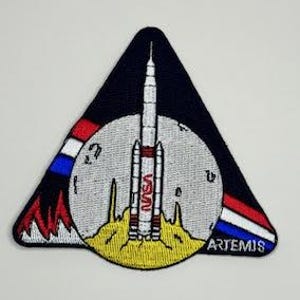 May include: A triangular patch with a red, white, and blue stripe design. The patch features a rocket launching from a yellow surface with a gray moon in the background. The text "ARTEMIS" is printed in white at the bottom of the patch. The text "NASA" is printed in red on the side of the rocket.