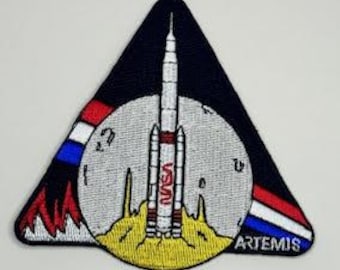 May include: A triangular patch with a red, white, and blue stripe design. The patch features a rocket launching from a yellow surface with a gray moon in the background. The text "ARTEMIS" is printed in white at the bottom of the patch. The text "NASA" is printed in red on the side of the rocket.