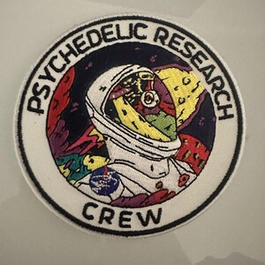 Può includere: Una toppa circolare bianca con testo ricamato nero che recita "PSYCHEDELIC RESEARCH CREW". Il centro della toppa presenta un astronauta colorato con un casco, circondato da una scena spaziale psichedelica.